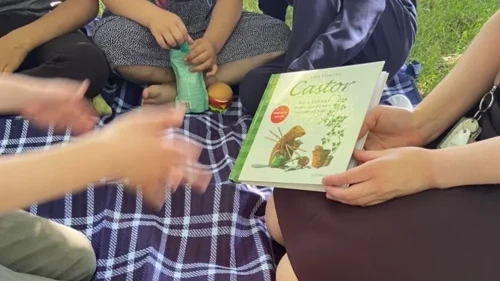En grupp med barn sitter  på en filt tillsammans med en  person som håller i en barnbok